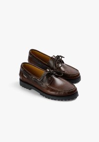 Zapatos de barco de cuero marrón con punta redondeada, cordones y suela de goma negra contrastante. Presentan costuras decorativas y un logo lateral.