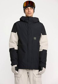 Veste imperméable noire et gris clair avec capuche, logo sur la poitrine et poches latérales. Portée avec des gants, associée à un pantalon beige.