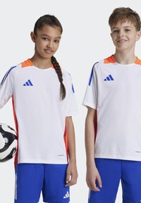 Két gyermek sportöltözetet visel, amely fehér pólóból áll, narancssárga és kék díszítéssel, valamint kék rövidnadrágból. Az egyikük egy labdát fog a kezében.