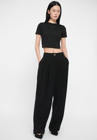 Γυναίκα φορώντας ένα μαύρο κοντό μανίκι μπλουζάκι και ψηλόμεσο μαύρο παντελόνι σε φαρδιά γραμμή, με τα χέρια στις τσέπες, μπροστά σε άσπρο φόντο.