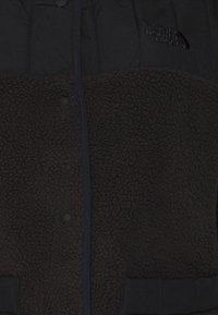 Giacca in pile nera con una parte inferiore testurizzata, pannello superiore liscio, bottoni a pressione e un logo sul petto.