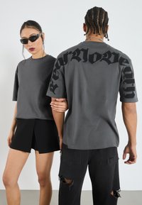 Grå kortärmade t-shirts har djärva svarta bokstäver på ryggen. Den manliga modellen bär slitna svarta jeans; den kvinnliga modellen matchar med svarta shorts.