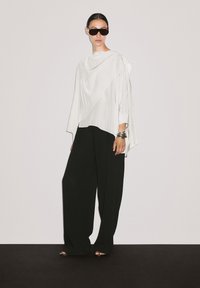 Haut blanc drapé avec une hem asymétrique ; associé à un pantalon large noir ample. Le mannequin porte des lunettes de soleil noires et des bracelets métalliques.