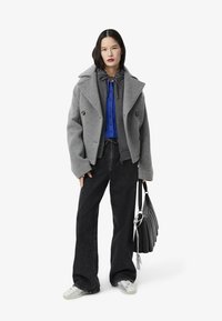 Cappotto grigio in lana a doppio petto con ampi rever, indossato sopra una camicia blu con bottoni, abbinato a jeans neri a gamba larga e una borsa nera.
