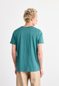 Camiseta de algodón de manga corta en color teal con cuello redondo, que presenta una textura suave y costuras mínimas, vista desde la parte de atrás, combinada con pantalones beige.