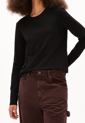 Woman wearing a black long-sleeve shirt tucked into high-waisted brown pants with a side loop on the right leg.