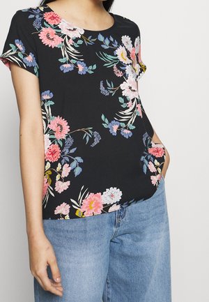 Kobieta w czarnej, krótki rękaw bluzce w kwiaty w kolorach różowym, białym i niebieskim, w zestawie z jasnoniebieskimi jeansami, z ręką w kieszeni.