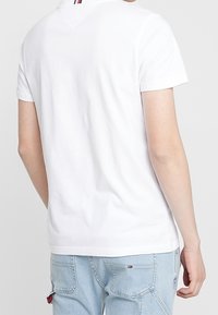 Camiseta blanca de manga corta con cuello redondo, que presenta un detalle a rayas en la parte posterior del cuello. La tela parece suave y lisa.