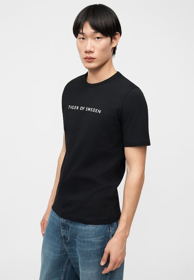 Νεαρός άνδρας που φοράει μαύρο T-shirt "Tiger of Sweden" και μπλε τζιν, στέκεται μπροστά σε απλό ανοιχτόφωτο φόντο.