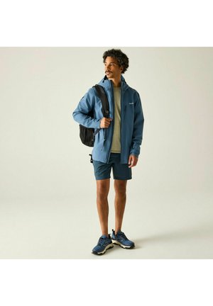 Man wearing blue jacket, gray shirt, blue shorts, and hiking shoes, carrying black backpack, standing on plain background looking to side.