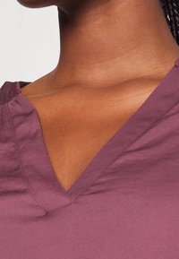 Top di cotone viola con scollo a V, cuciture visibili e texture liscia. Il tessuto ha una leggera lucentezza e una vestibilità comoda.