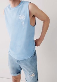 Chemise sans manches bleu clair avec un ourlet arrondi et des broderies de palmier. Portée avec un short en denim délavé, avec des revers roulés.