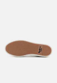 Semelle d'une chaussure en caoutchouc marron avec des rainures diagonales et un bord blanc, présentant un patch logo rectangulaire près du talon.