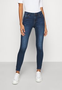 Tynne mørkeblå jeans med middels høy livhøyde, stilige sømmer og subtil slitasje ved knærne, kombinert med hvite joggesko.