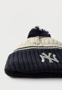 Pletene kapa z rebrasto teksturo v mornarsko modri in krem barvi, z cofom na vrhu in vezenim logotipom NY spredaj.