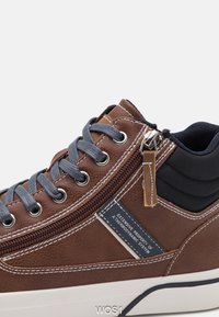 Bruna syntetiska lädersneakers med grå snören, blixtlåsdetalj, vadderad svart krage, vit sömnad och en etikett med text på sidan.