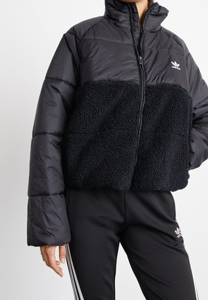 Personne portant une doudoune noire Adidas avec un panneau en polaire et un pantalon Adidas noir assorti avec des bandes blanches.