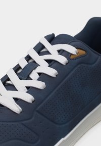 Gros plan sur une basket bleu marine avec des lacets blancs, des panneaux latéraux perforés et un détail œillet beige sur une semelle blanche.