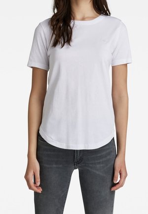 Kvinde iført en ensfarvet hvid T-shirt med korte ærmer og mørkegrå jeans, stående med armene afslappet langs siden mod en hvid baggrund.