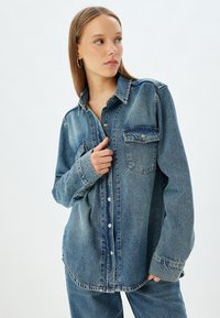 Camisa de mezclilla en azul claro con mangas largas, cierre frontal de botones, un bolsillo en el pecho y con dobladillo ligeramente curvado. La tela parece duradera y texturizada.