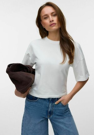 Donna con lunghi capelli castani che indossa una maglietta bianca a maniche corte e jeans blu, tenendo una giacca marrone scuro piegata su un braccio.