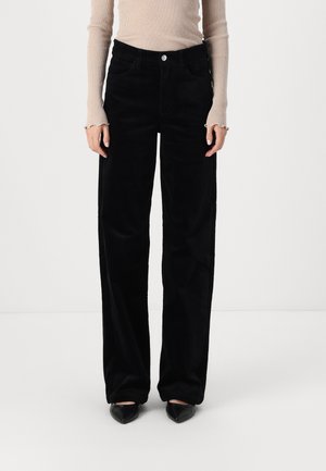 Femme portant un pantalon noir à jambes larges, un haut beige à manches longues côtelé et des chaussures noires à bouts pointus, debout devant un fond blanc uni.