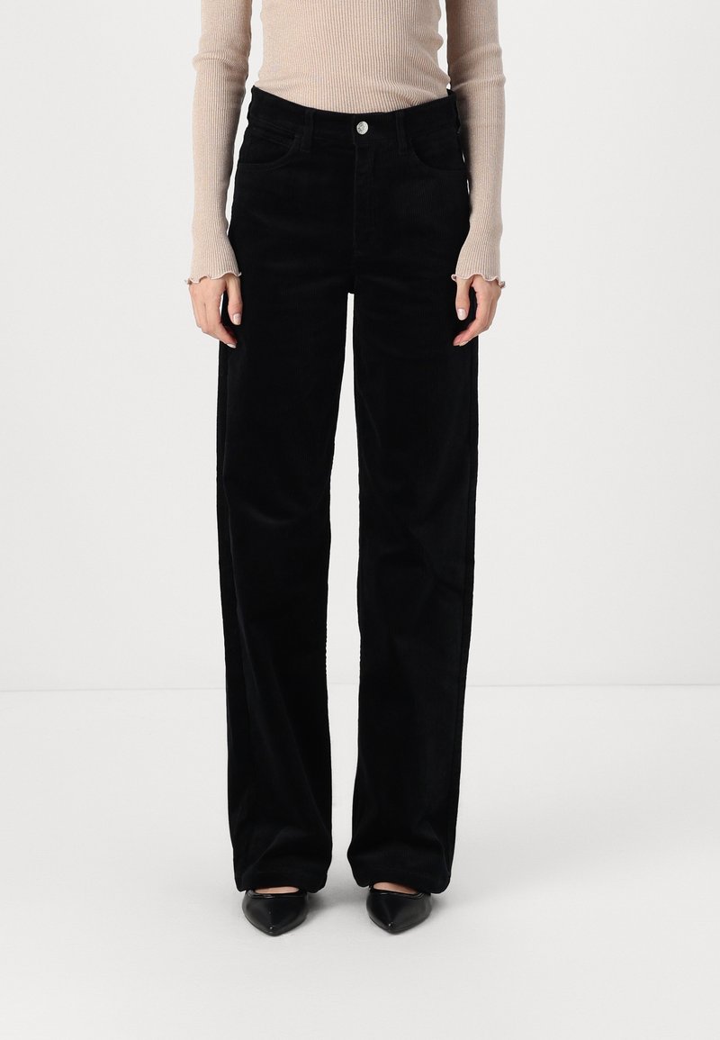 Femme portant un pantalon noir à jambes larges, un haut beige à manches longues côtelé et des chaussures noires à bouts pointus, debout devant un fond blanc uni.