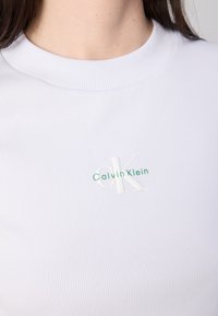 Close-up van een persoon die een geribbeld, wit T-shirt draagt met een groen "Calvin Klein" logo en een gestileerde "CK" op de borst.