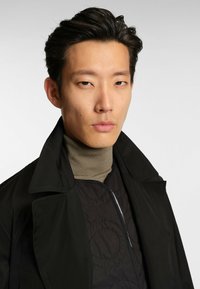 Czarny płaszcz z szerokim kołnierzem, nałożony na fakturowaną ciemną kurtkę z okrągłymi wzorami, noszony na jasnobrązowym golfie.