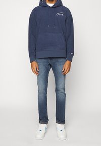 Navyblauwe hoodie met een voorzak, verstelbare trekkoord en wit logo, gecombineerd met lichtblauwe jeans en witte sneakers.