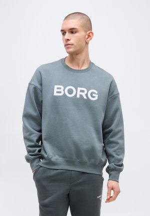 Fiatal férfi rövid hajjal, szürke pulóvert visel, amelyen a "BORG" felirat olvasható, oldalra néz, kezei a nadrág zsebeiben.