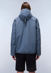 Chaqueta impermeable azul claro con capucha ajustable, cierre delantero y corte relajado, llevada sobre pantalones cortos negros.