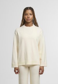 Maglione lavorato a maglia color crema con collo rotondo, spalle scese e polsini a coste. La texture morbida e la vestibilità rilassata si abbinano ai pantaloni coordinati.