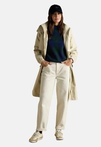 Bēša vējjakas ar aukliņu, tumši zaļš argyle džemperis, krēmkrāsas džinsi un bēši sporta apavi. Cepure ir tumši zila ar logo.