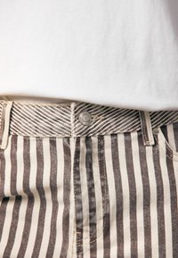 Jean en denim rayé beige et marron avec fermeture à bouton, passants de ceinture et tissu texturé. Caractéristiques des coutures visibles et un motif distinct.