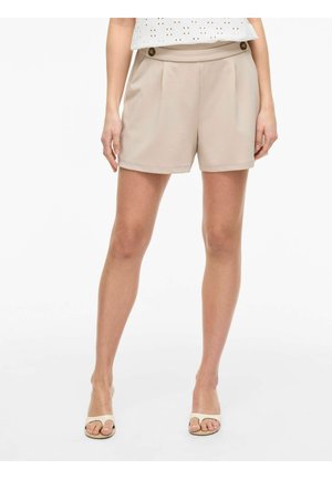 Femme portant un short beige taille haute avec plis devant et détails de boutons, associé à des talons ouverts blancs et un haut blanc.