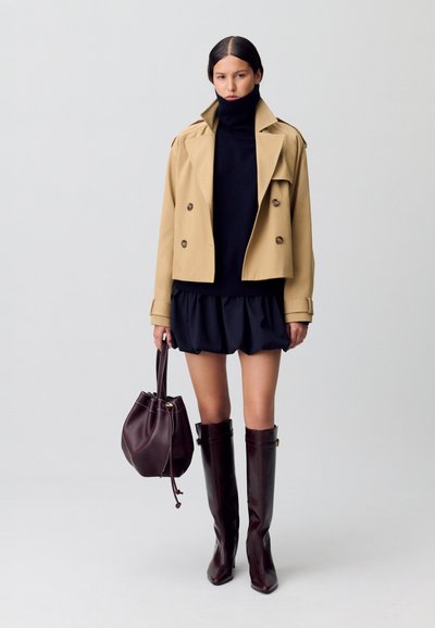 Femme portant un trench-coat court beige, un pull noir à col roulé, une mini-jupe noire, des bottes bordeaux montantes, tenant un sac à main bordeaux.