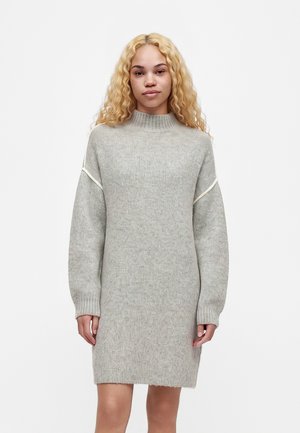 Jeune femme aux longs cheveux blonds bouclés portant une robe-pull en tricot gris clair à manches longues, debout devant un fond blanc.