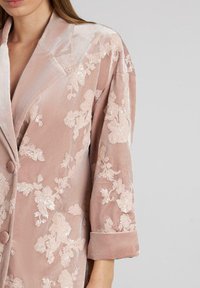 Blazer de terciopelo rosa empolvado con bordados florales, solapas estructuradas y mangas enrolladas; textura suave con detalles de lentejuelas.