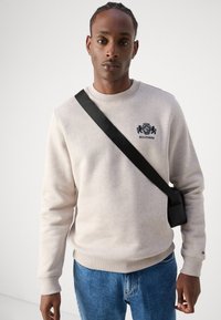 Lys grå sweatshirt med ribbede manchetter og nederkant, forsynet med et marineblåt broderet logo. Sammen med blå jeans og en sort crossover taske.