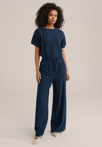 Outfit blu navy con una maglietta morbida e pantaloni a gamba larga, realizzati in tessuto soffice. Presenta una vita con cintura e scarpe basse con dettagli squadrati.