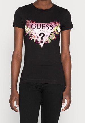 Sort kortærmet tætsiddende t-shirt med pink glitrende GUESS-logo, blomstermotiv og kædedesign, båret af en person med sorte bukser.