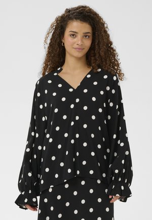 Moteris su garbanotais plaukais, vilkinti juodą ilgomis rankovėmis palaidinę su baltais taškeliais ir derančią sijoną, stovinti priešais paprastą foną.