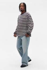 Ungdom med flettehår iført svart-hvite striper langermet skjorte, lyseblå baggy jeans, svarte sko, står mot hvit bakgrunn.