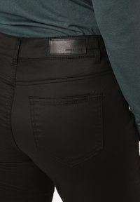 Schwarze, enganliegende Jeans mit glatter Textur, ausgestattet mit Gesäßtaschen und einem Leder-Label. Das Oberteil ist türkis und teilweise über dem Bund sichtbar.