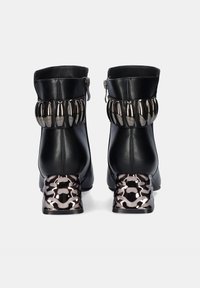 Botas de tobillo de cuero negro con una banda metálica decorativa y tacones de bloque con patrón. Presentan un cierre de cremallera lateral.