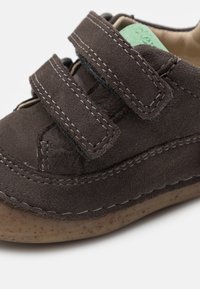 Zapato de ante marrón oscuro con dos correas de Velcro, detalles de costura clara, forma redondeada y suela de goma moteada.