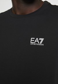 Černé bavlněné tričko s kulatým výstřihem a krátkými rukávy. Na levé straně hrudi je bílé vyšívané logo "EA7 EMPORIO ARMANI".