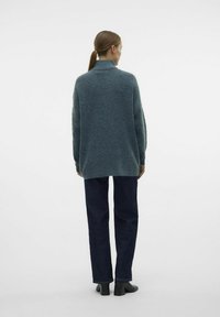 Prevelik teal pulover z visokim ovratnikom, izdelan iz teksturiranega kvačkanega materiala. Ujemajoč s temnimi jeans hlačami in črnimi gležnjarji.