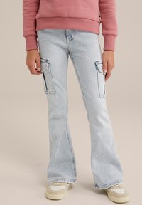 Světlé rozšířené džíny s kapsami ve stylu cargo na stehnech, prošívanými detaily a měkkou texturou, doplněné béžovými a krémovými teniskami.